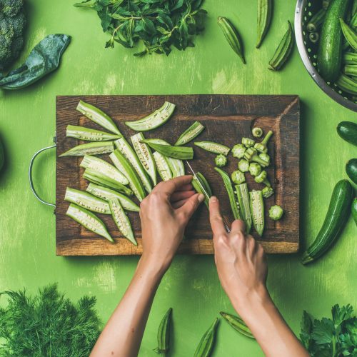 Healthy green vegan cooking ingredients. Flay-lay of female hands cutting green vegetables and greens over green background, top view. Clean eating, vegetarian, detox, dieting food concept